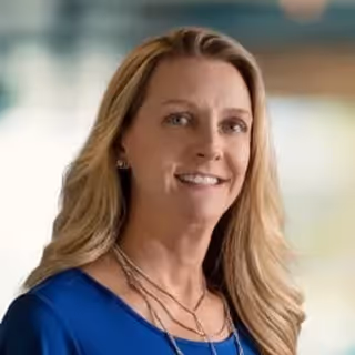 Smiling woman with long blonde hair wearing a blue top and layered necklaces against a blurred background.