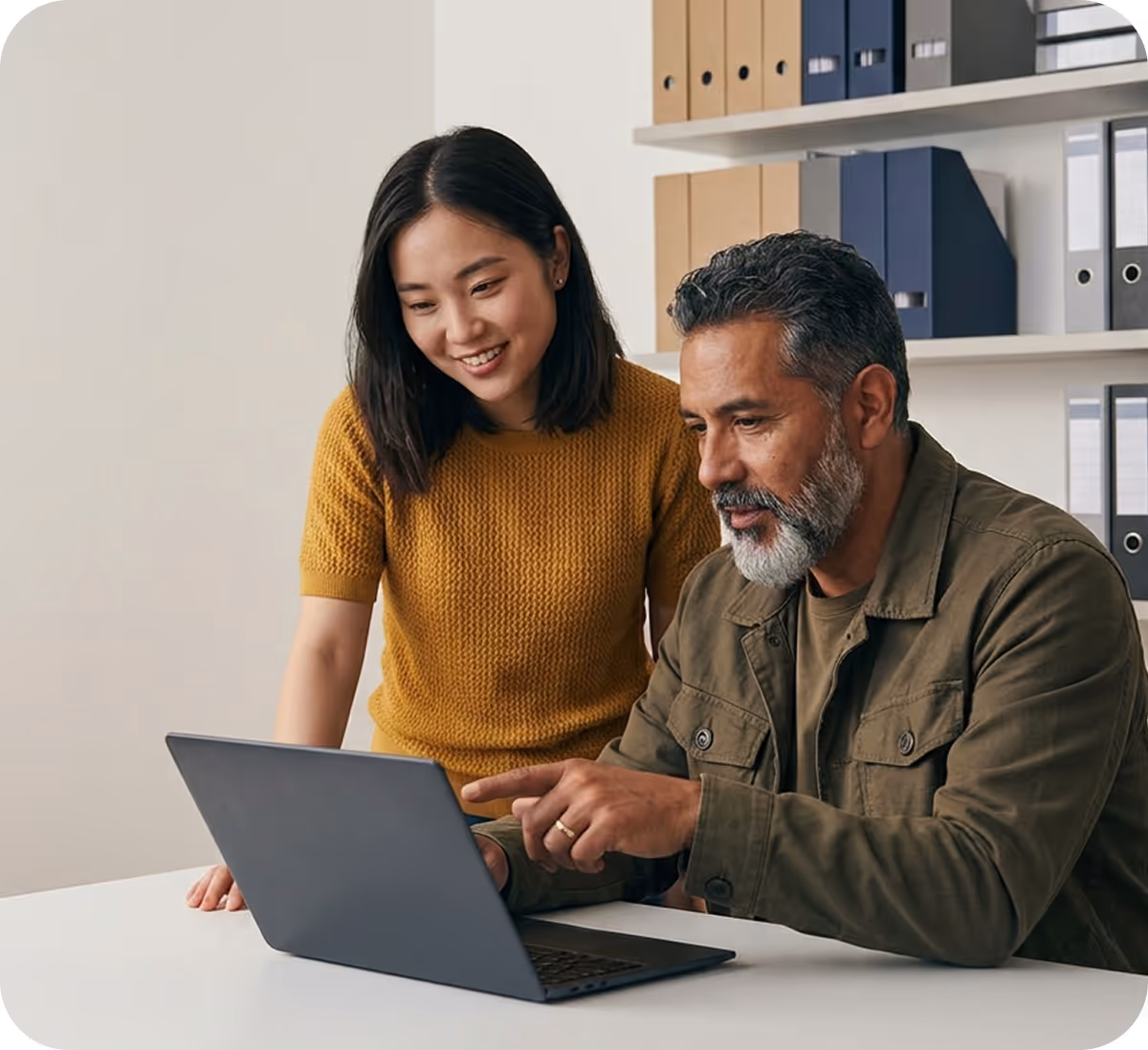 Man and woman looking at a laptop screen together in an office setting with shelves of files in the background.