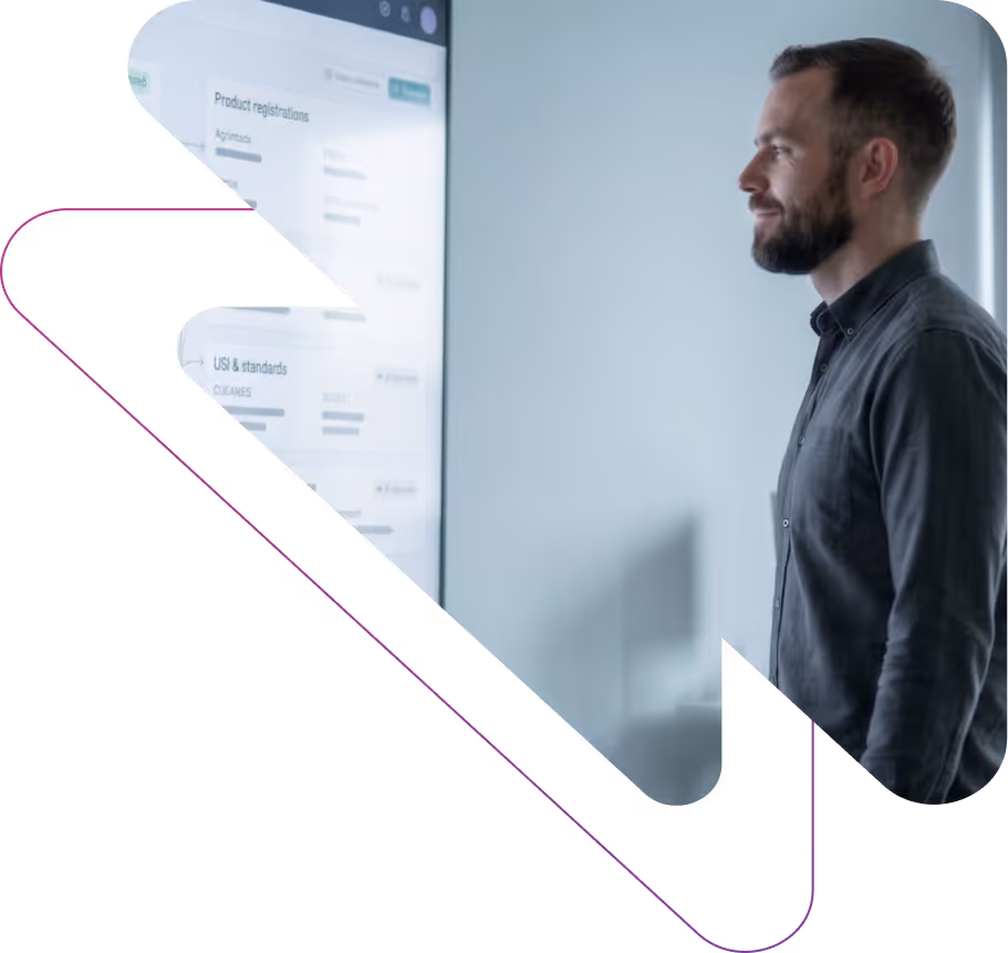 A man with a beard and dark shirt looking at a large screen displaying product registrations and standards information in a modern office.