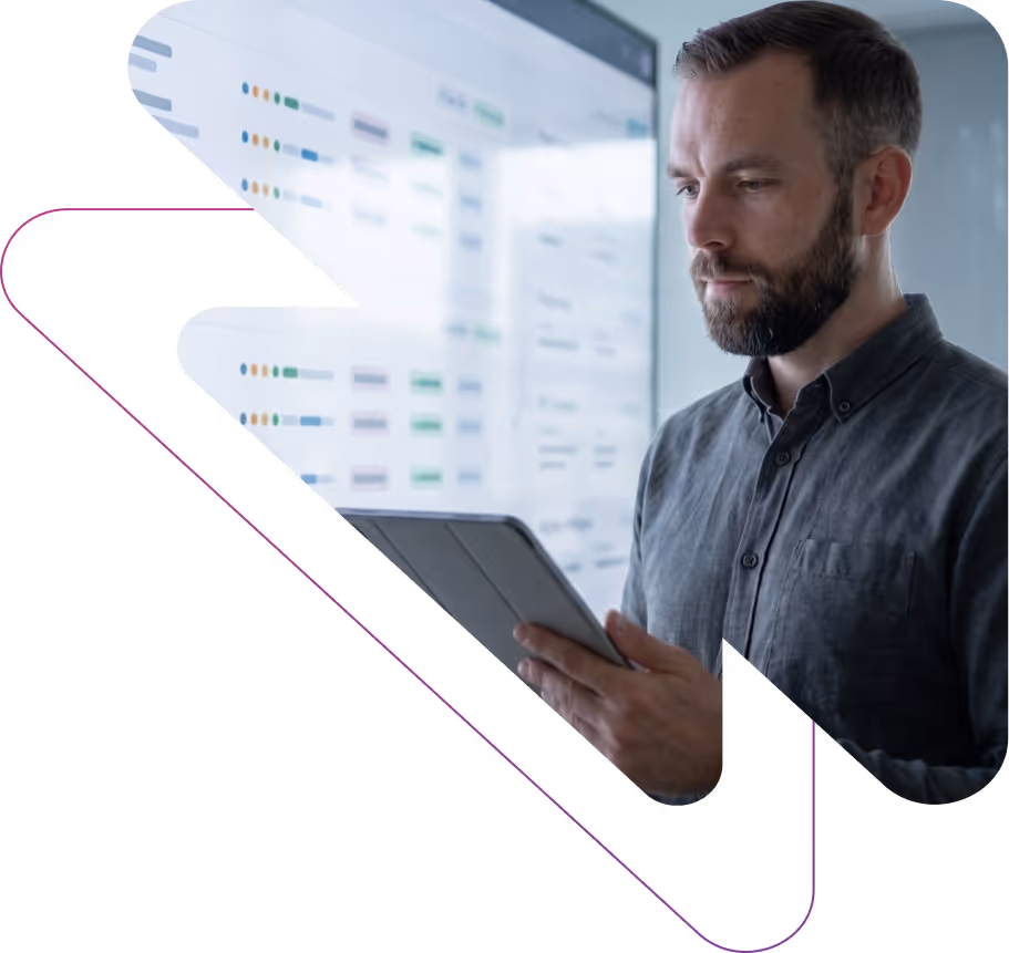 Man with beard holding a tablet and looking at a large screen displaying data charts and tables.