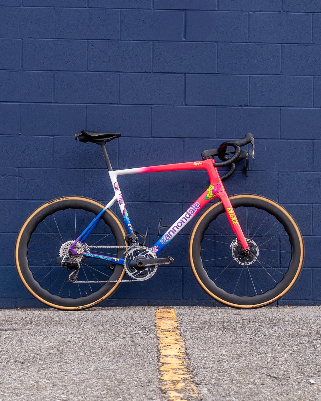 Cannondale road bicycle with pink, white, and blue frame and tan tire sidewalls against a blue brick wall.