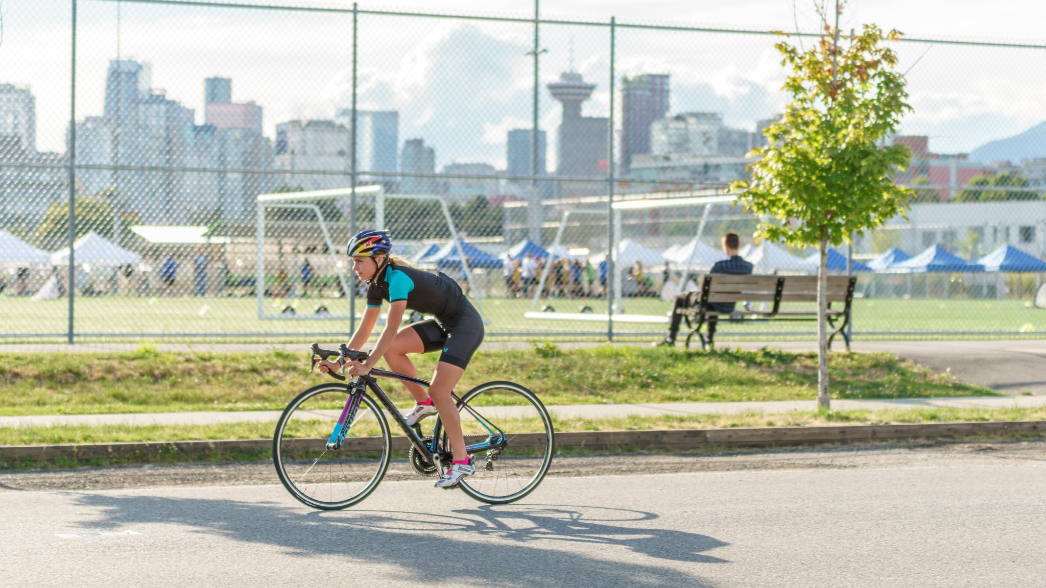 Person in cycling gear riding a road bike on a street near a fenced soccer field with a cityscape in the background.