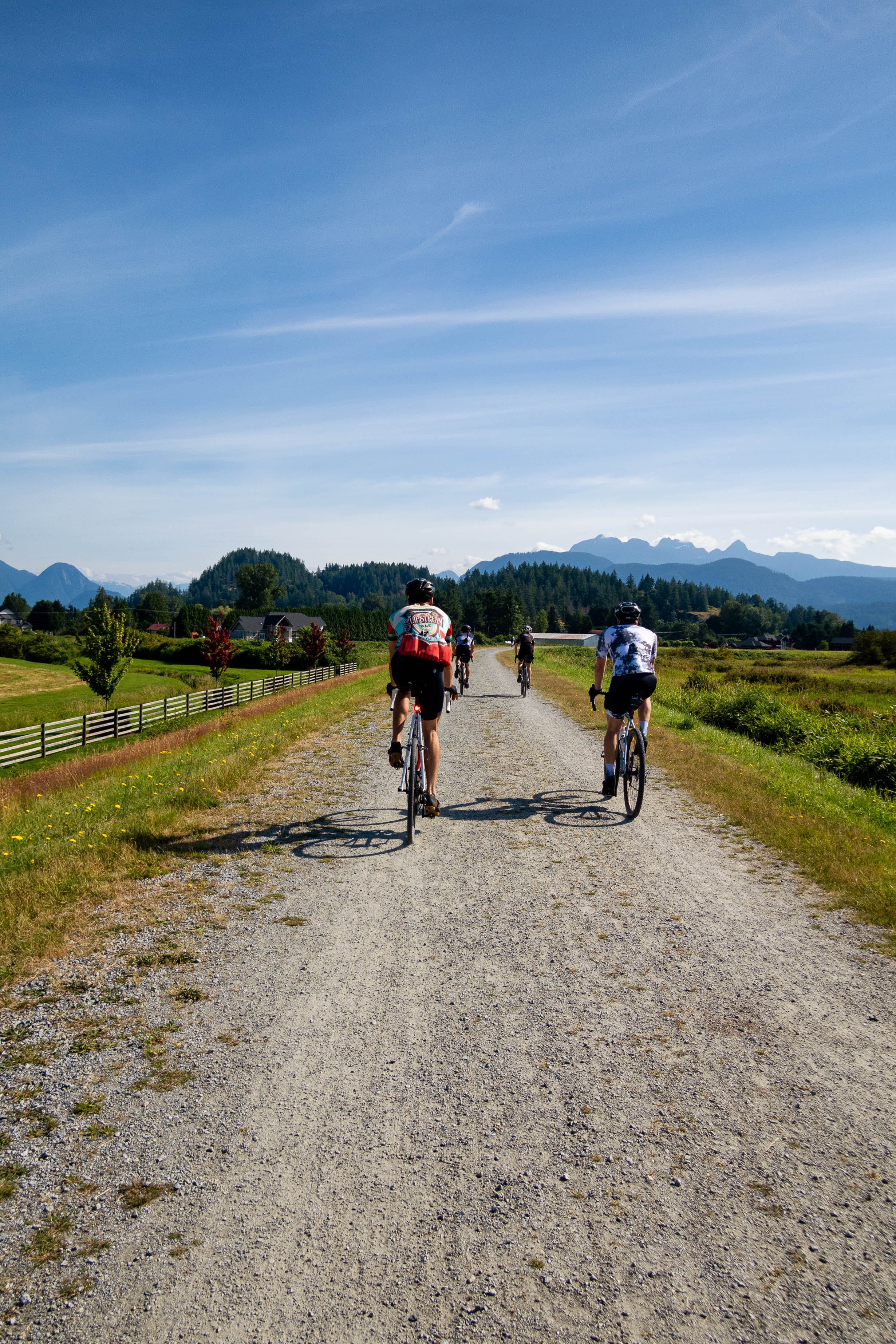 Group of cyclists riding on a gravel path through a green rural landscape with mountains in the background under a clear blue sky.