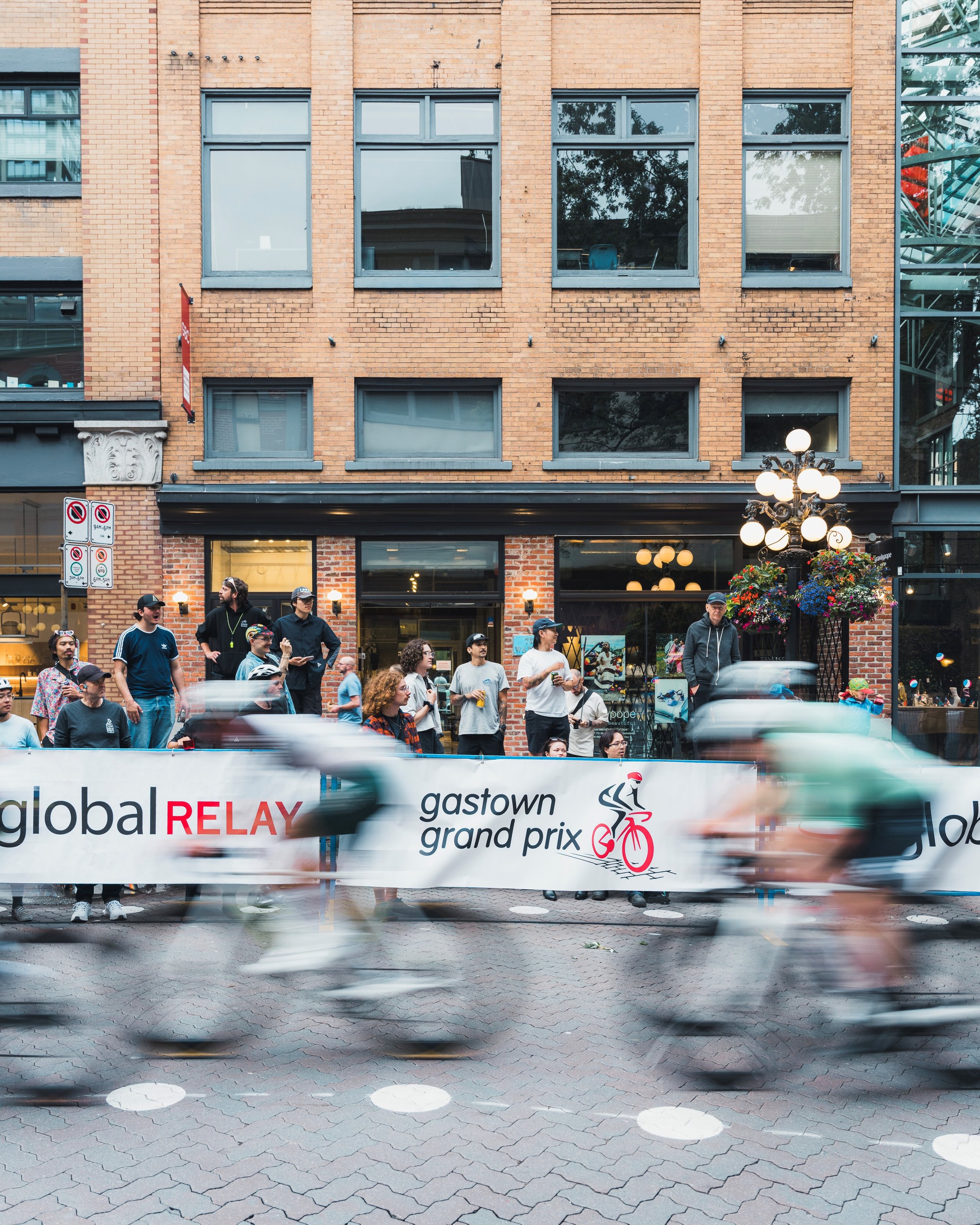 Motion-blurred cyclists racing past spectators along a street with a Gastown Grand Prix banner in front of a brick building.