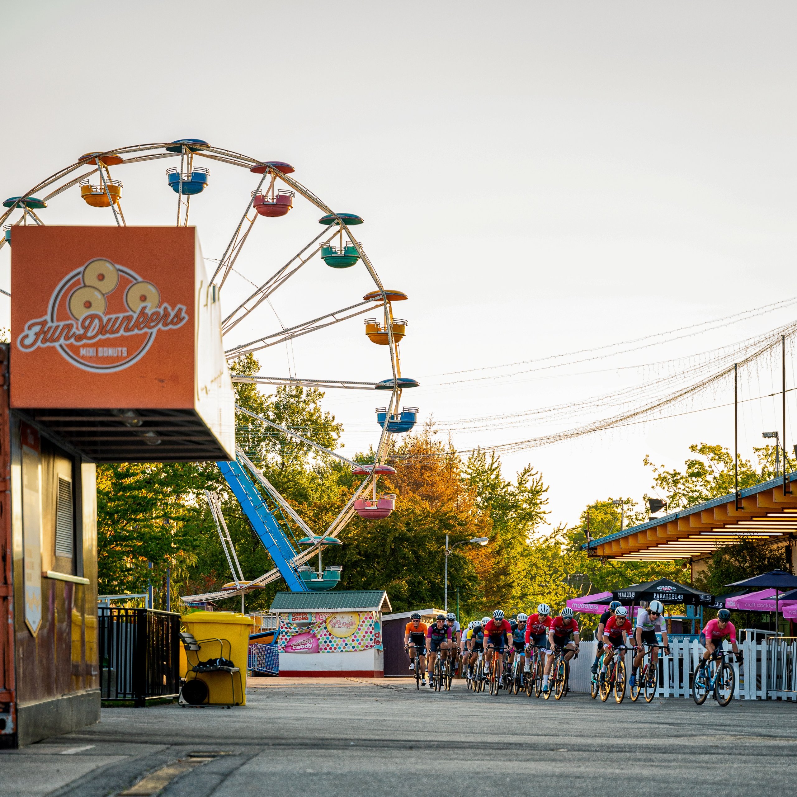 Group of cyclists racing on a paved path near a colorful Ferris wheel and food stands during daylight.