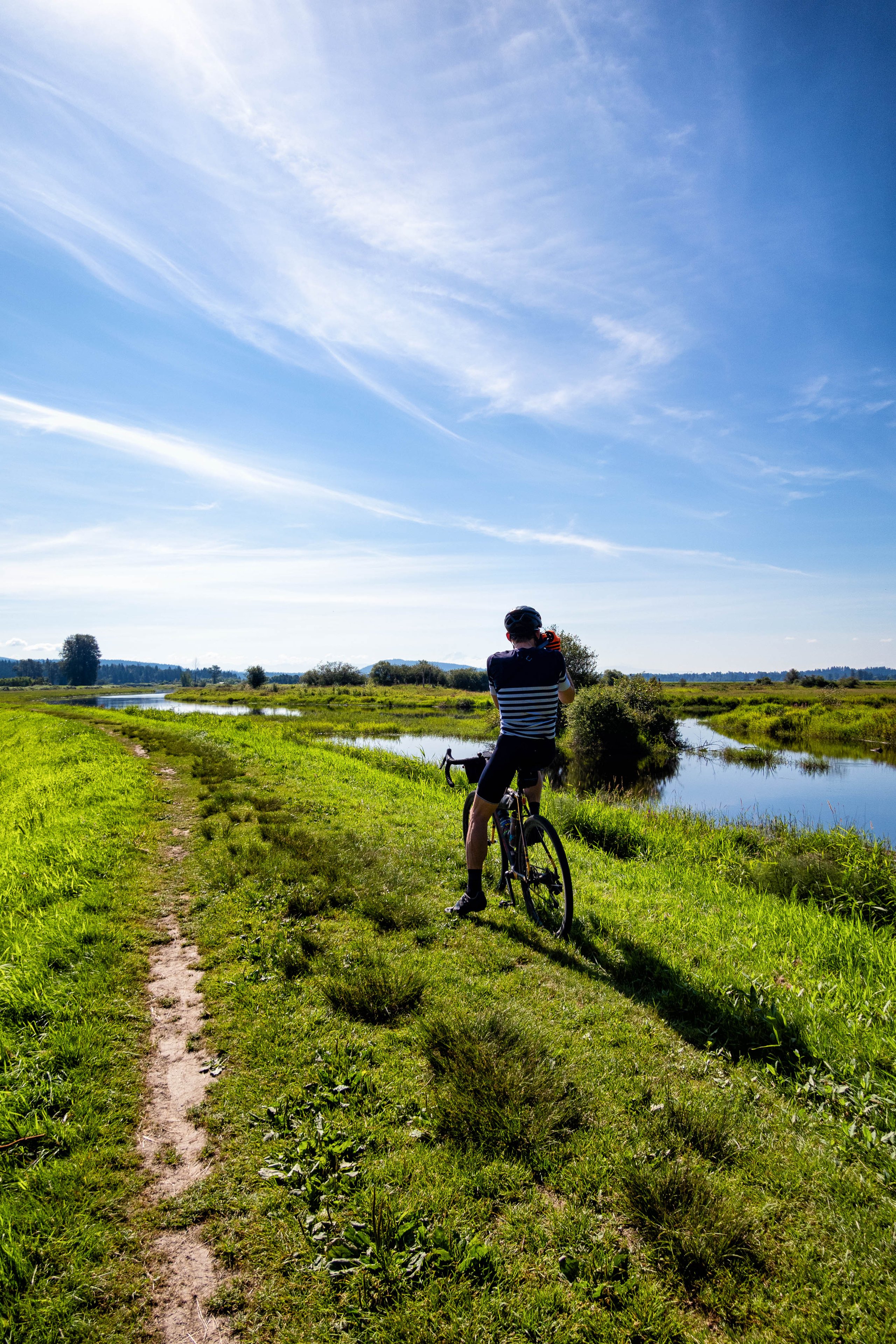 Cyclist in helmet and striped jersey standing on grassy riverside path, taking a photo with a camera under a blue sky with wispy clouds.