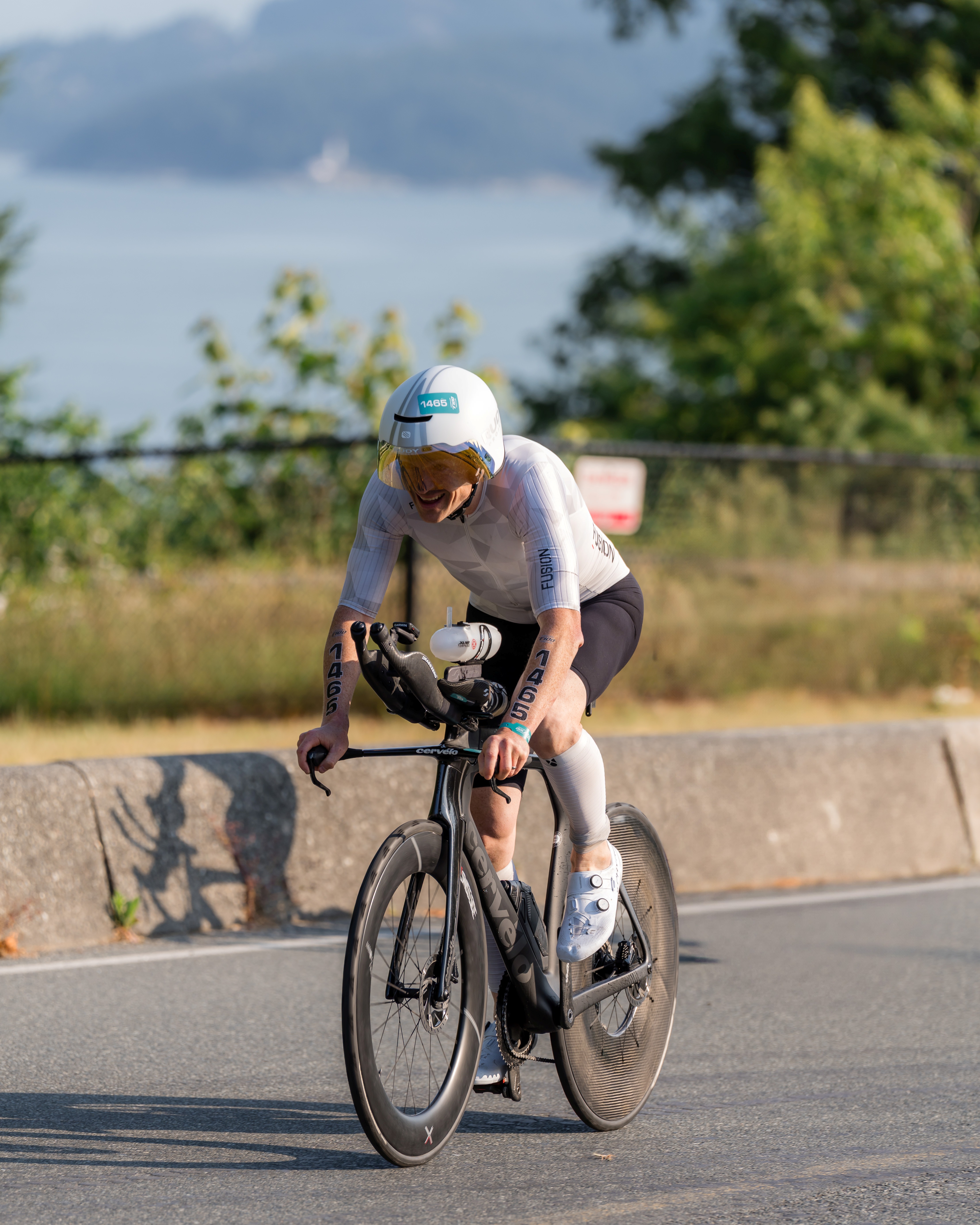 Cyclist wearing white aerodynamic helmet and gear riding a black Cervelo time trial bike on a road with greenery and water in the background.