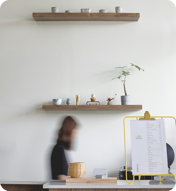 Minimalist interior with two wooden shelves holding small pottery, a potted plant, and a blurred person standing behind a counter next to a menu on a yellow stand.