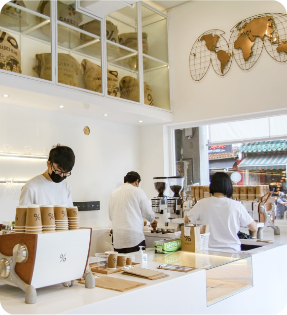 Three baristas working behind a white counter in a modern coffee shop with stacks of paper cups and coffee machines.