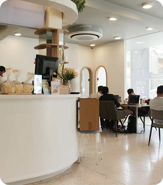 Modern café interior with a counter displaying packaged bread, plants, and a computer monitor, alongside customers seated at tables with laptops.