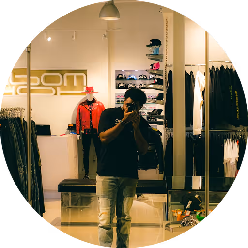 Man taking a photo of himself in a clothing store mirror with racks of clothes and mannequins in the background.
