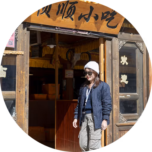 Woman wearing sunglasses, a white beanie, and a blue jacket standing outside a wooden building with Chinese characters above the entrance.