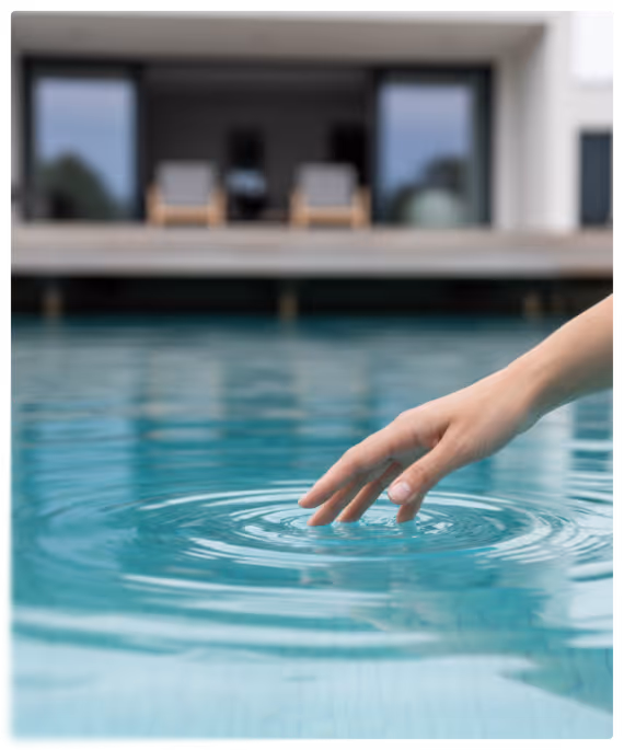 Une main touchant doucement la surface bleue d'une piscine créant des ondulations, avec une maison moderne floue en arrière-plan.