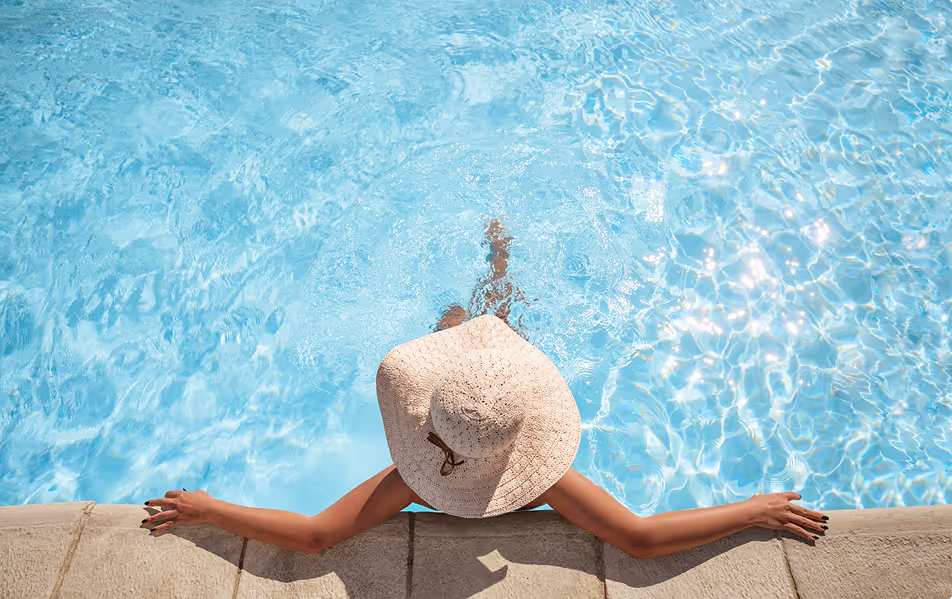 Personne vue de dessus dans une piscine bleue, portant un large chapeau beige et tenant les bords de la piscine.