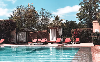 Bord d'une piscine avec des transats rouges et une cabane ombragée entourée d'arbres sous un ciel bleu partiellement nuageux.