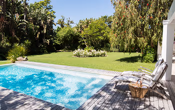 Piscine à débordement avec eau bleue et fauteuils en bois sur une terrasse en bois, entourée d'un jardin verdoyant sous un ciel dégagé.