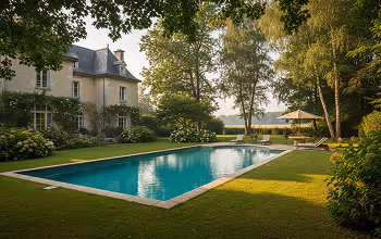 Piscine rectangulaire entourée d'une pelouse verte devant une grande maison beige avec un toit mansardé, arbres et parasol à l'arrière.