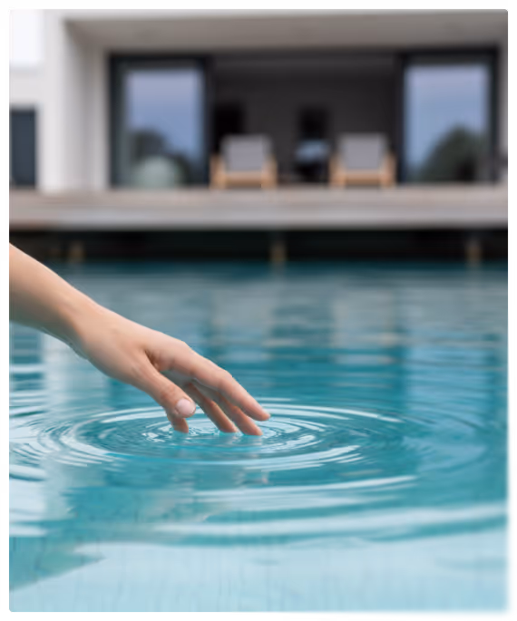Main touchant délicatement la surface de l'eau d'une piscine devant une maison moderne avec des fauteuils sur la terrasse.