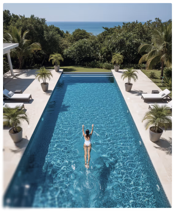 Femme en maillot de bain blanc nageant dans une piscine rectangulaire avec des palmiers en pots autour, vue sur la mer et des arbres en arrière-plan.