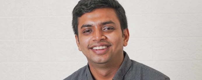 Smiling man with short black hair wearing a dark patterned shirt, against a light textured background.