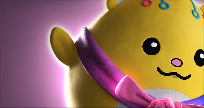 Close-up of a yellow plush toy with big round eyes, a smiling mouth, and a purple and pink scarf on a purple glowing background.