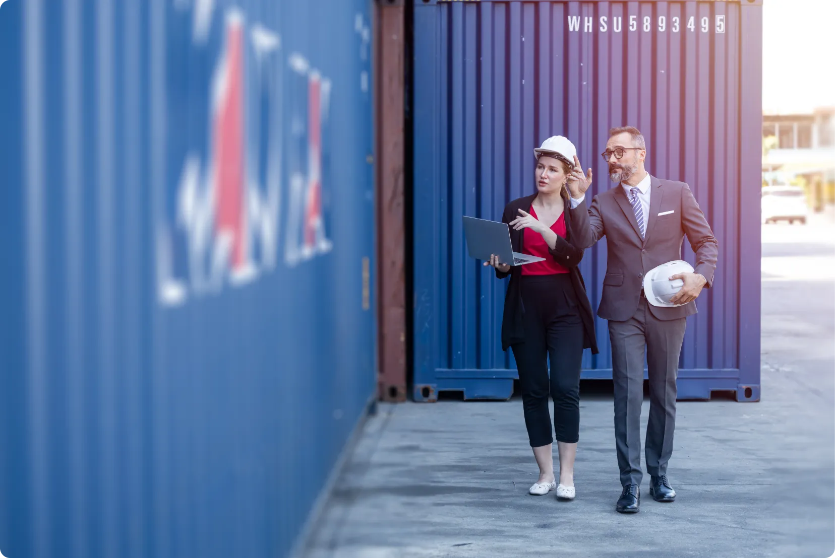 Man and woman wearing hard hats inspecting blue shipping containers, woman holds a laptop.