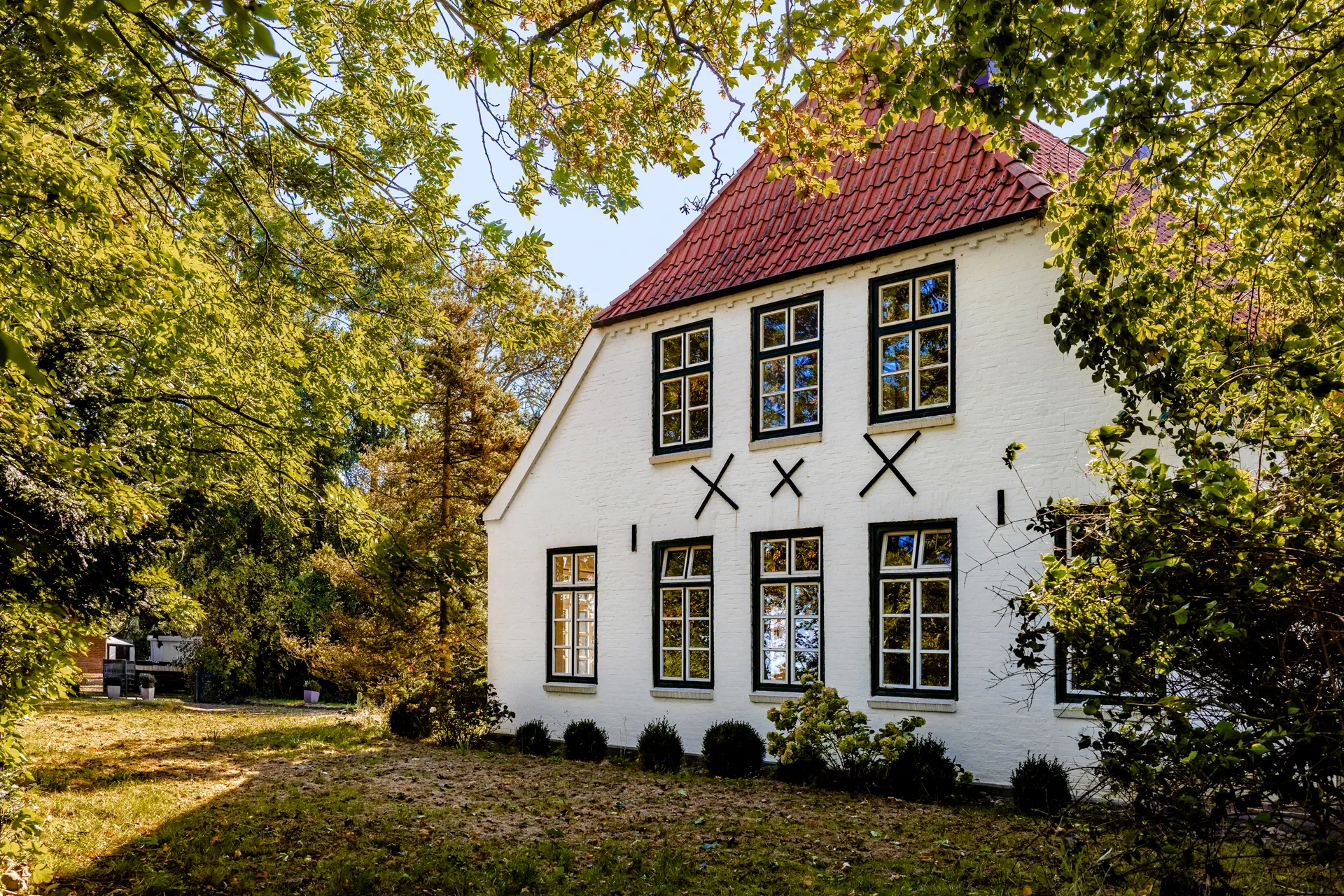Weißes Haus mit roten Ziegeldach und mehreren Fenstern, umgeben von grünem Garten und Bäumen.