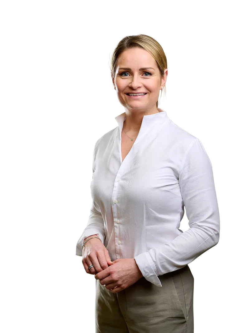 Lächelnde Frau mit blonden Haaren trägt weiße Bluse und beige Hose, vor schwarzem Hintergrund.