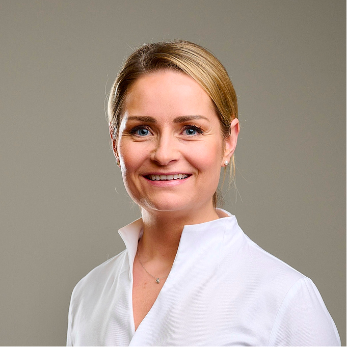 Porträt einer lächelnden blonden Frau mit blauen Augen in weißer Bluse vor grauem Hintergrund.