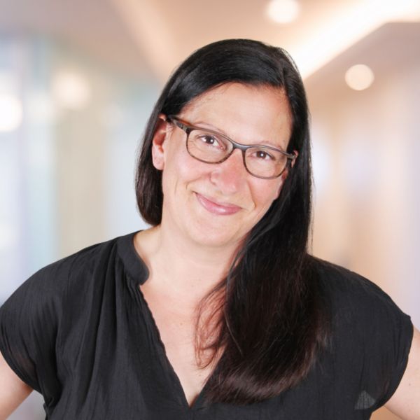 Lächelnde Frau mit langen dunklen Haaren und Brille vor unscharfem Hintergrund.