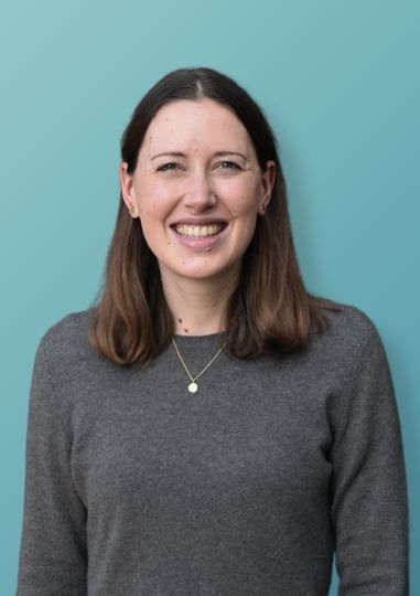 Junge Frau mit braunen schulterlangen Haaren, grauem Pullover und kleinem goldenen Halsanhänger, die vor einem hellblauen Hintergrund lächelt.