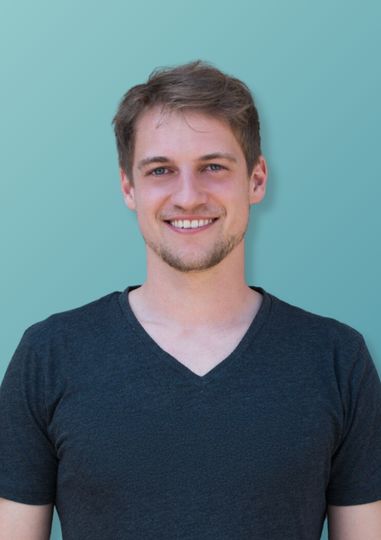 Junger Mann mit kurzem braunem Haar und schwarzem V-Ausschnitt-T-Shirt lächelt vor hellem blaugrünem Hintergrund.