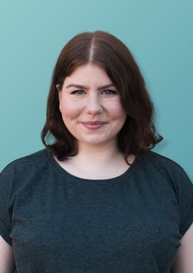 Porträt einer lächelnden Frau mit schulterlangen braunen Haaren vor einem hellblauen Hintergrund.