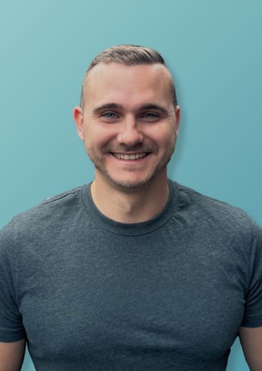 Lächelnder Mann mit kurzen blonden Haaren und grauem T-Shirt vor einem hellblauen Hintergrund.