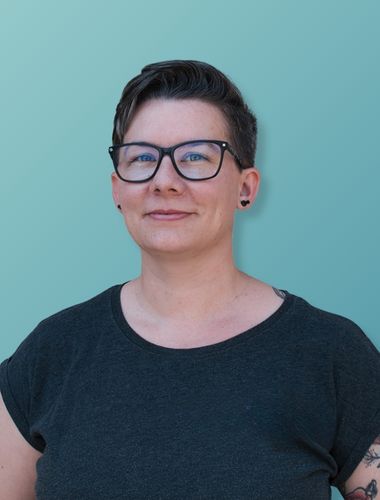 Portrait einer Person mit kurzen, dunklen Haaren, schwarzen Brillengestell und dunklem T-Shirt vor hellblauem Hintergrund.