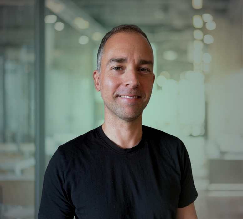 Porträt eines lächelnden Mannes mit kurzem Haar und schwarzem T-Shirt vor einem modernen Bürohintergrund.