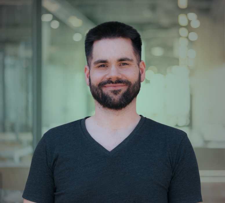 Porträt eines lächelnden Mannes mit dunklem Haar und Bart, der ein dunkelgraues T-Shirt trägt, vor einem verschwommenen Bürohintergrund.