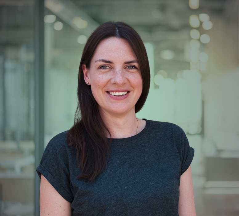 Porträt einer lächelnden Frau mit langen braunen Haaren, die ein dunkelgraues T-Shirt trägt, vor einem unscharfen Bürohintergrund.