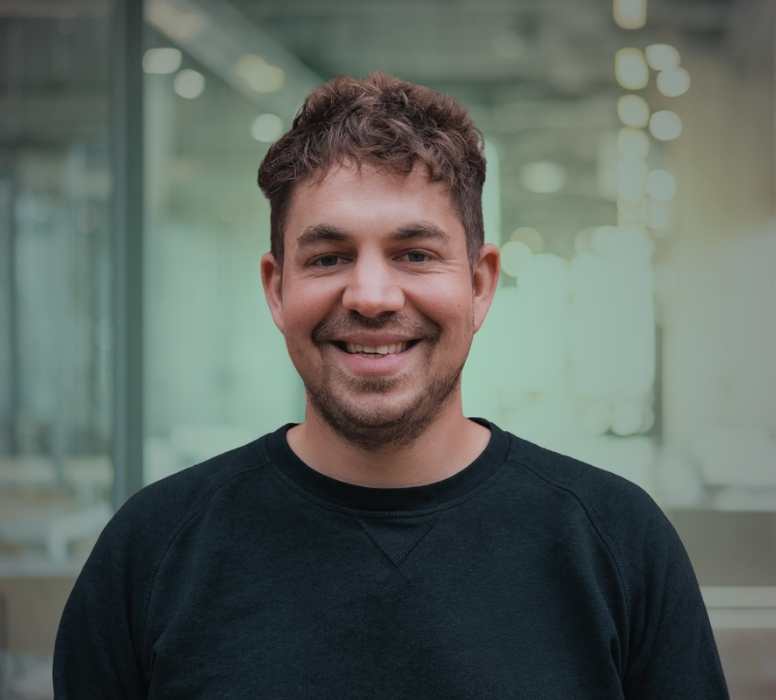 Porträt eines lächelnden Mannes mit kurzen lockigen Haaren und dunklem Pullover vor einem verschwommenen Bürohintergrund.