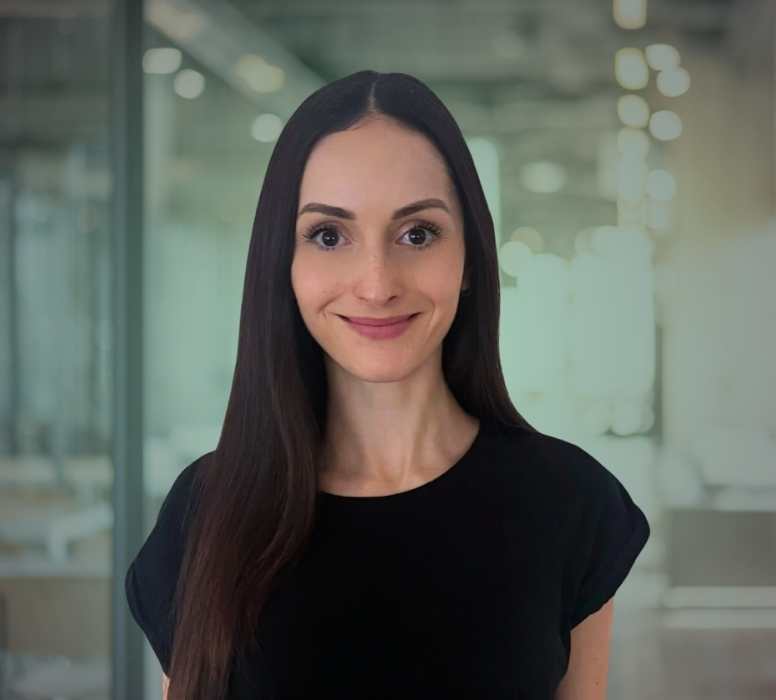 Porträt einer lächelnden Frau mit langen, glatten dunklen Haaren vor einem modernen Bürohintergrund.