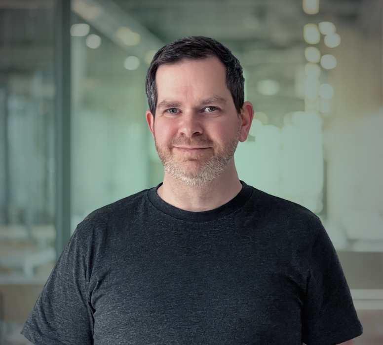 Porträt eines lächelnden Mannes mit kurzen dunkelbraunen Haaren und leichtem Bart, der ein dunkles T-Shirt trägt und vor einem unscharfen modernen Bürohintergrund steht.