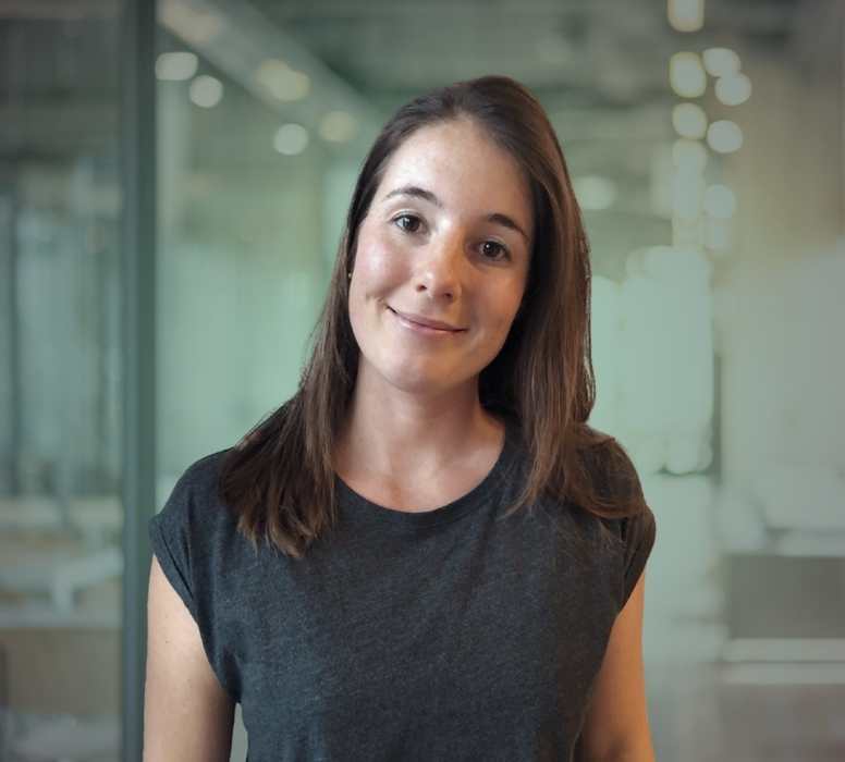 Porträt einer lächelnden Frau mit braunen Haaren in einem dunklen T-Shirt vor einem Bürohintergrund.