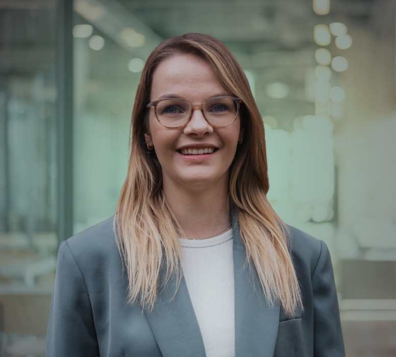 Junge Frau mit blonden Haaren, Brille und grauem Blazer lächelt in einem modernen Büro.