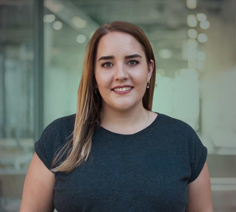 Porträt einer lächelnden jungen Frau mit langem, braunem Haar und dunklem T-Shirt vor einem verschwommenen Bürohintergrund.