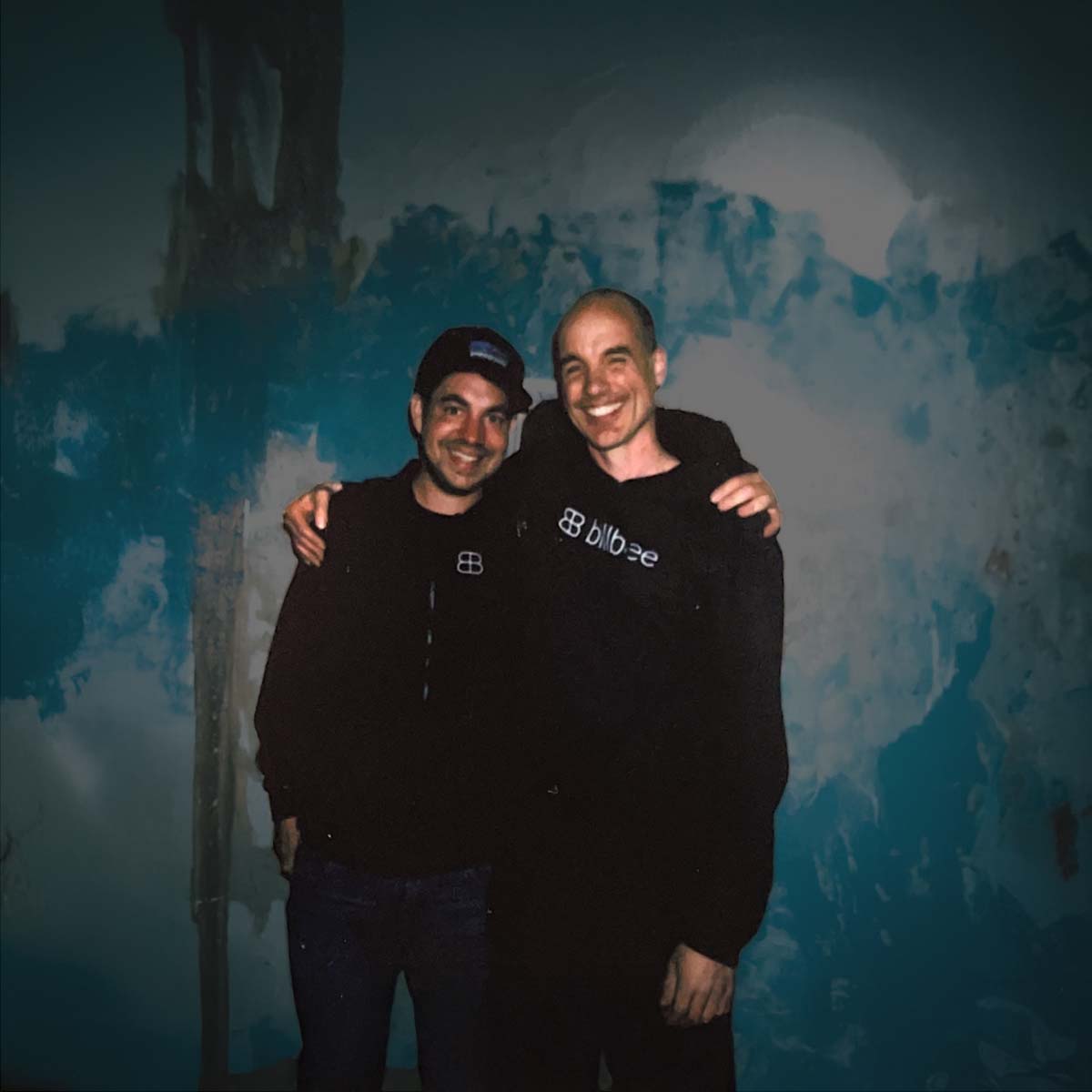 Zwei Männer lächeln und umarmen sich vor einer blau und weiß gestrichenen Wand.