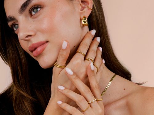 Frau mit makelloser Haut trägt goldene Ohrringe, Halskette und mehrere filigrane goldene Ringe an den Fingern vor beigem Hintergrund.