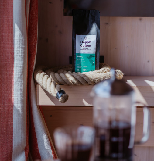 Kaffeepackung von Happy Coffee steht auf einem Holzbrett mit einer dicken Seilrolle, davor eine unscharfe French-Press-Kaffeekanne und -glas.