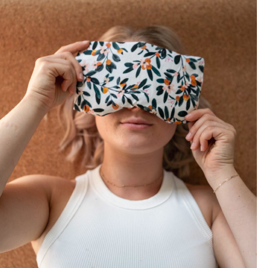 Frau hält eine Schlafmaske mit floralem Muster vor ihre Augen, vor braunem Hintergrund.