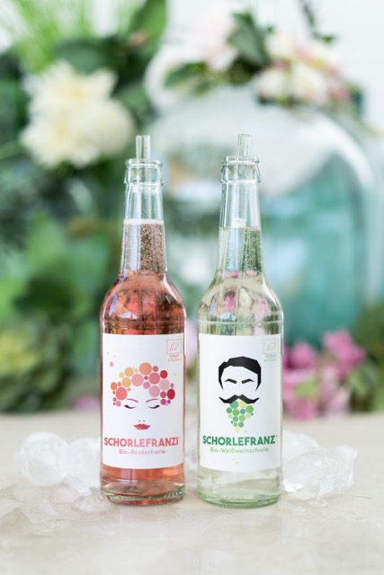 Zwei Flaschen Schorlefranzi Bio-Schorle, eine rosé und eine weiß, auf Eiswürfeln vor verschwommenem Blumenhintergrund.