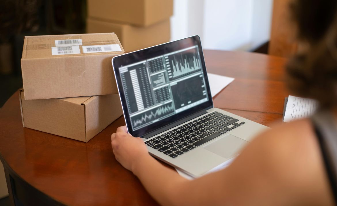 Eine Person sitzt an einem Tisch und betrachtet auf einem Laptop Datenvisualisierungen mit Diagrammen und Zahlen. Neben dem Laptop stehen zwei braune Versandkartons.